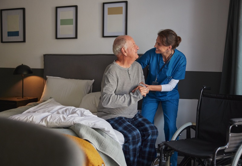 Caring nurse helping an elderly man in pajamas get up from bed in a bedroom. Mid adult healthcare professional supporting senior man while getting up from bed and move towards wheelchair at home. Compassionate caregiver providing support to old man during morning routine